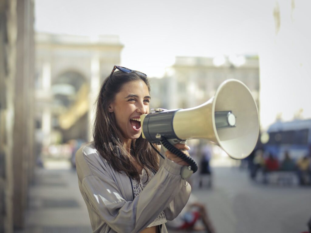 Cheerful young woman screaming into megaphone - employee benefits